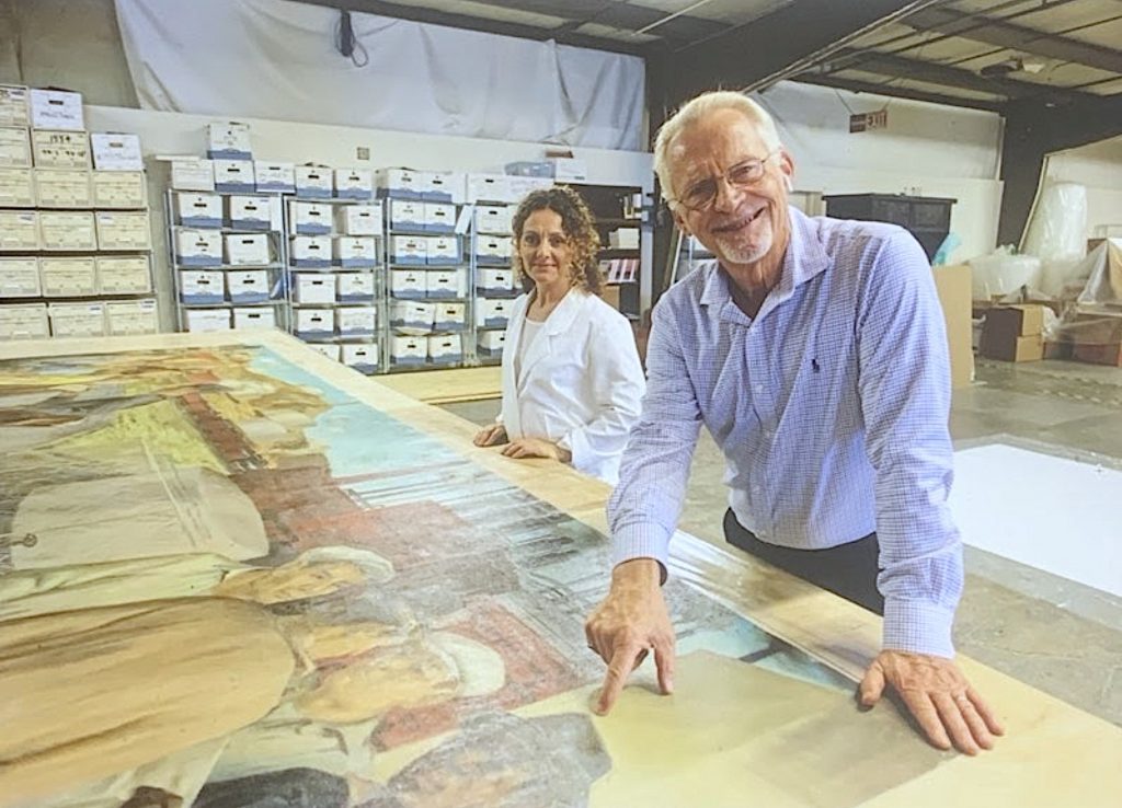 image of a mature white man standing next to a middle aged white woman as they reference a large painting splayed out on a table in front of them. They are FACL art conservators.